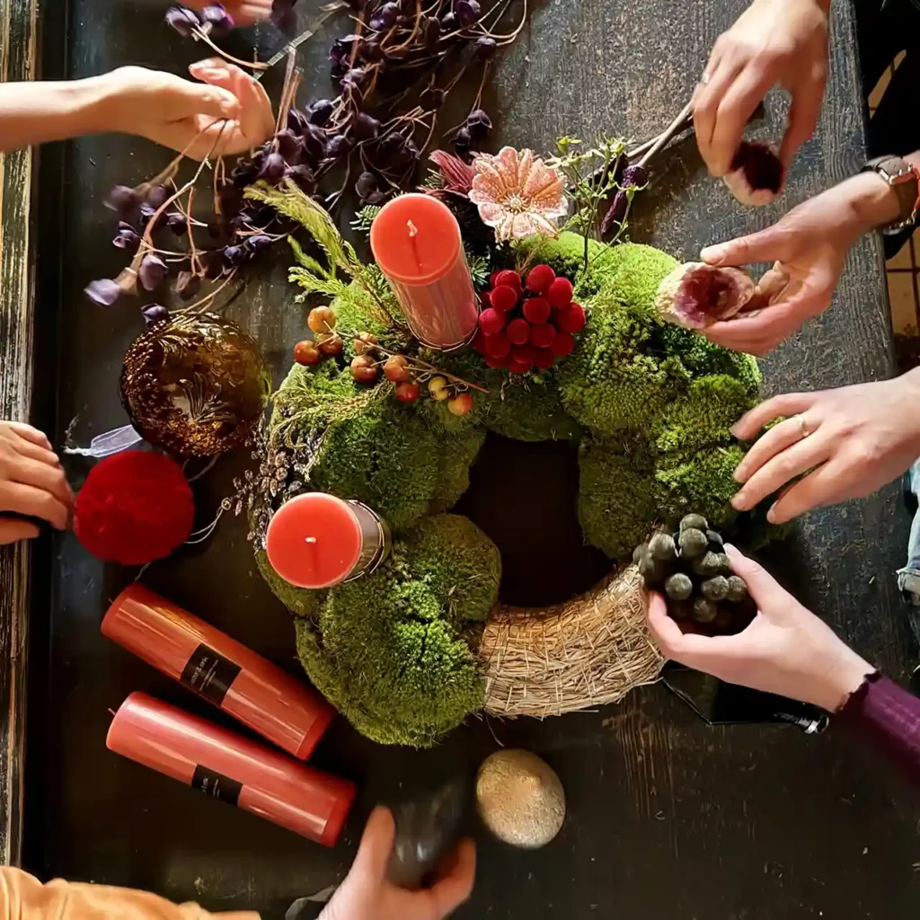 Mehrere Hände dekorieren einen außergewöhnlichen Adventskranz aus Moos mit roten Kerzen, Beeren und Naturmaterialien.