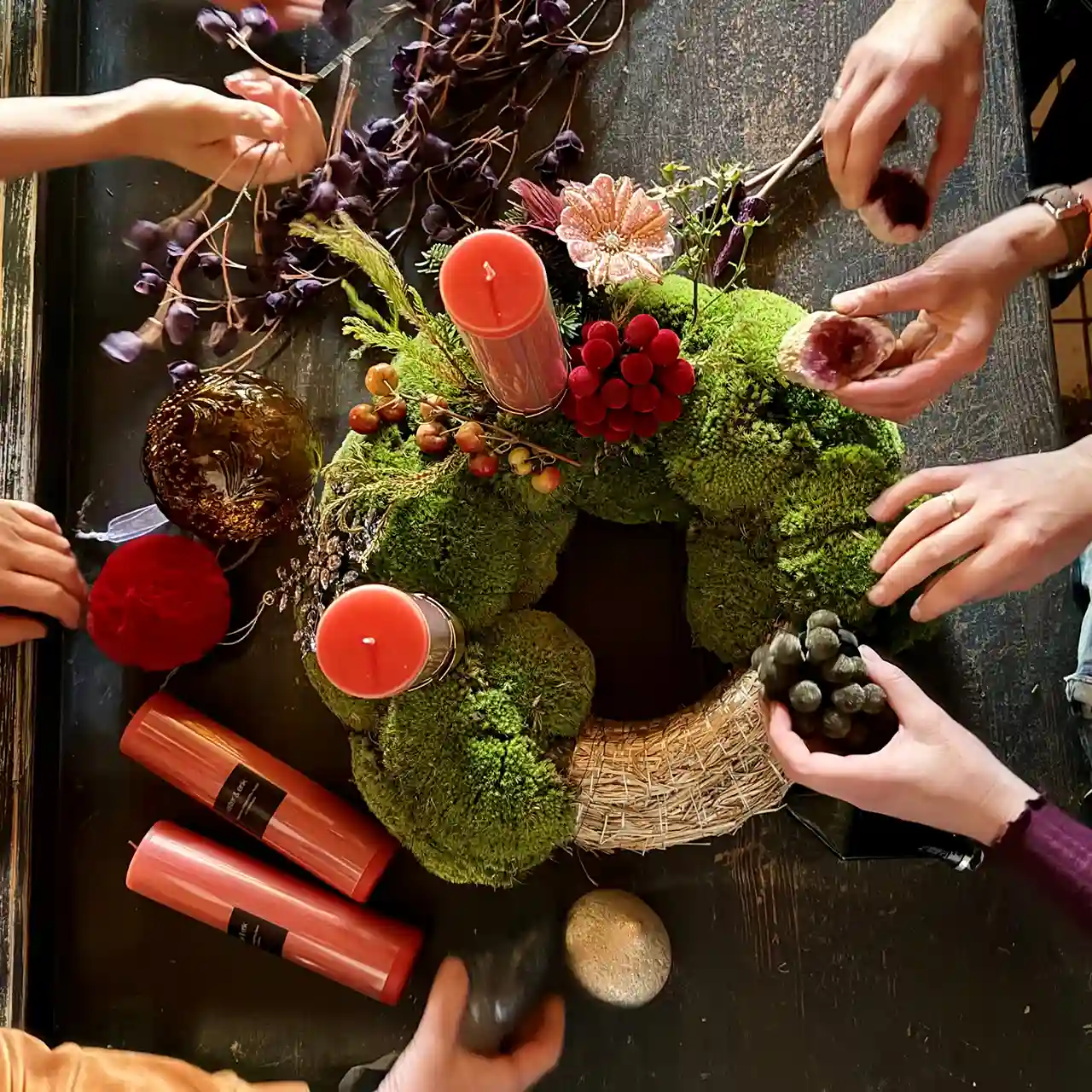 Mehrere Hände dekorieren einen außergewöhnlichen Adventskranz aus Moos mit roten Kerzen, Beeren und Naturmaterialien.